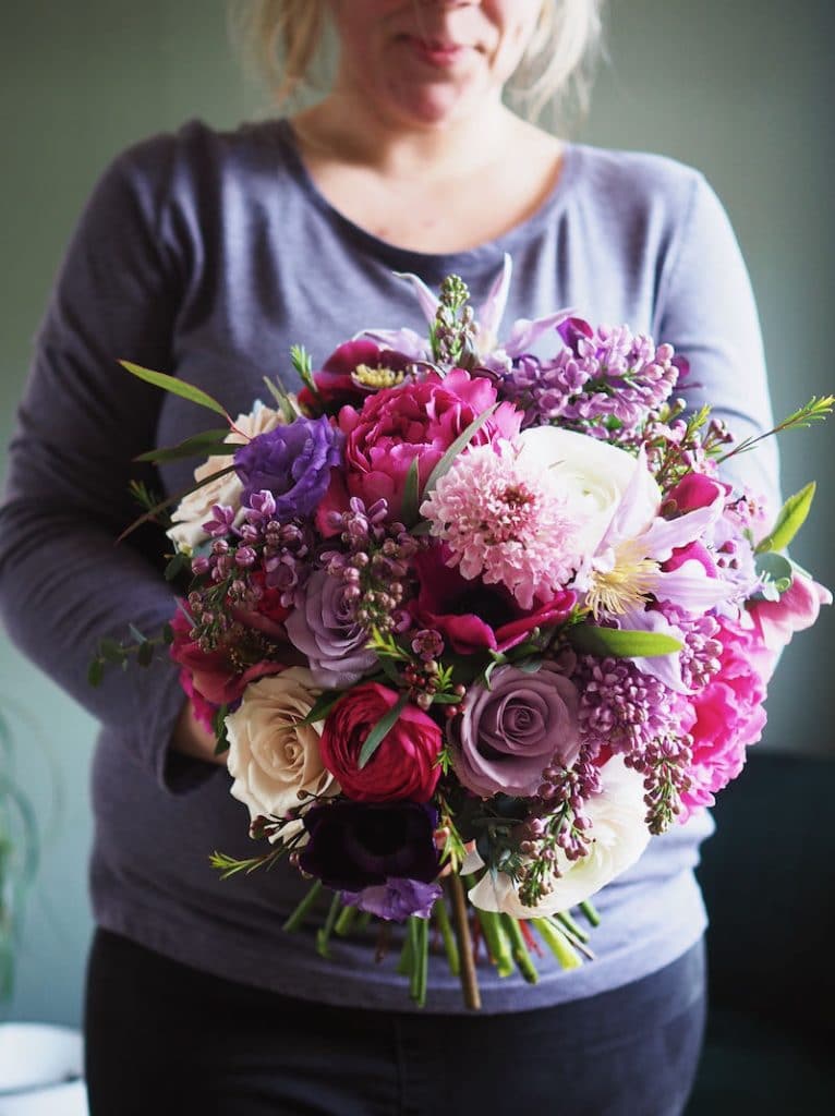 Image shows a florist holding a bright colourful spring wedding bouquet  at Farnham Castle. Roses, ranunculus, scabiosa, clematis and lilac are in the bouquet