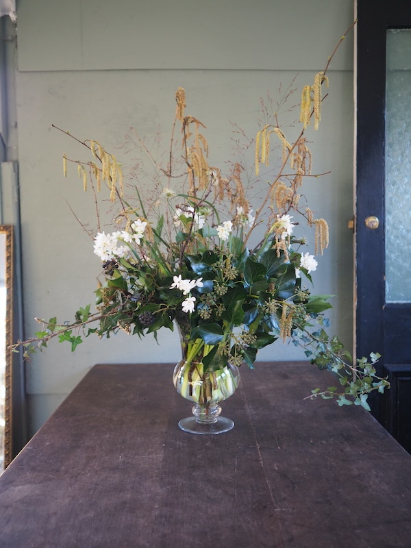 Image shows a large glass vase of winter flowers including catkins and paper whites. The vase is on a wooden table against a green background.