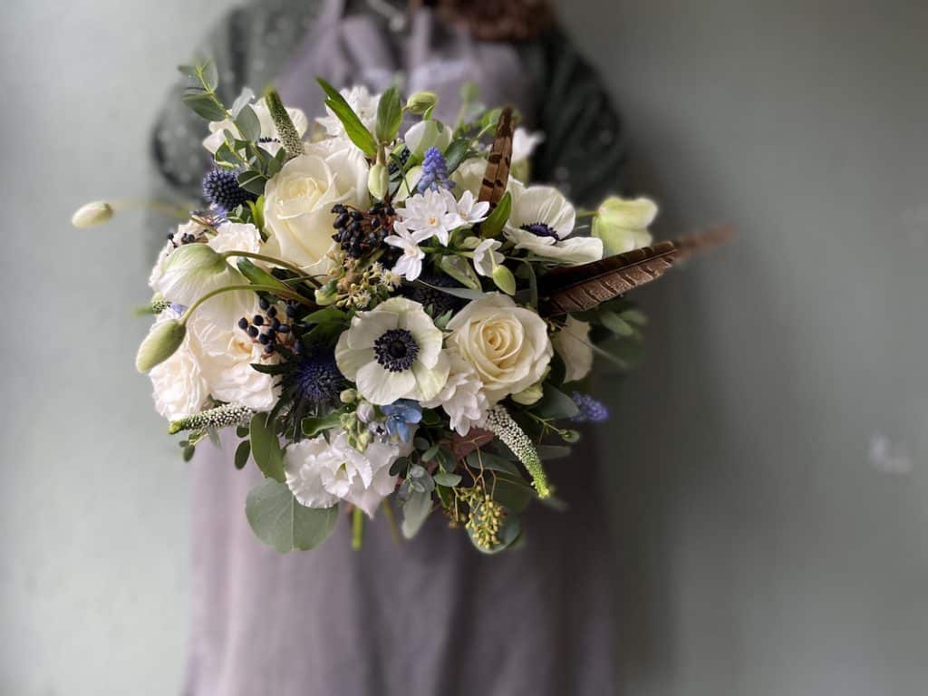 Picture shows a lady in a grey apron holding a bouquet of white flowers for a bride. The flowers are anemones and roses with pheasant feathers.