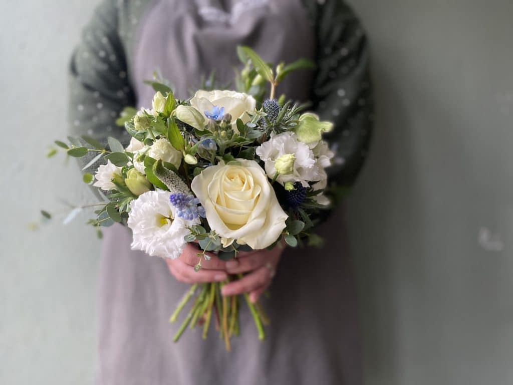 Picture shows a lady in a grey apron holding a bridesmaids bouquet. The bouquet contains white and blue flowers and is relaxed in design.