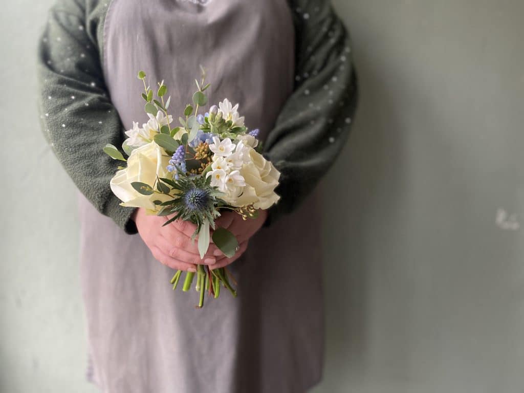 Picture shows a lady in a grey apron holding a small bouquet of flowers for a flower girl at a wedding. The flowers are white and blue.