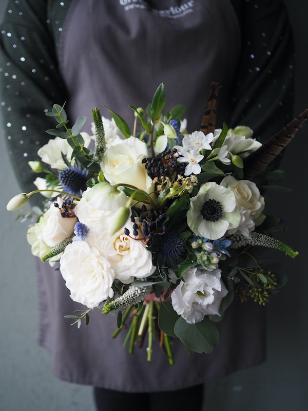 picture shows a lady in a grey apron holding a white bridal bouquet. The flowers are white, with berries and blue thistles