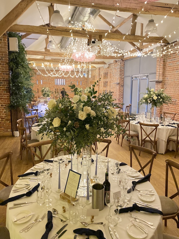 Picture shows a brick barn wedding venue with a large arrangement of flowers in a glass vase upon a table.