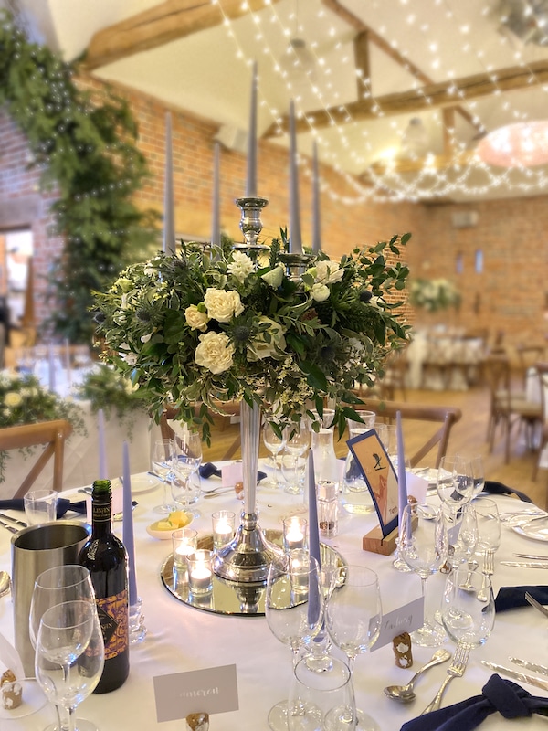 Picture shows a brick barn set up for a wedding. There are round tables and on the round tables there is a silver candelabra with a large arrangement of flowers on it.