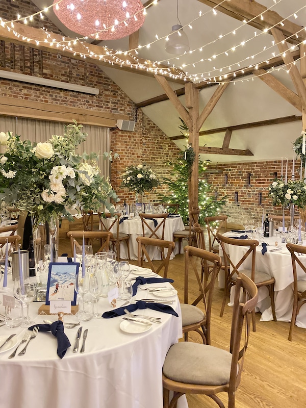 Picture shows an old brick barn set up for a wedding. There are round tables with white table cloths and on the tables there are large arrangements of white flowers. There are blue candles around the base and dark blue napkins on the tables.